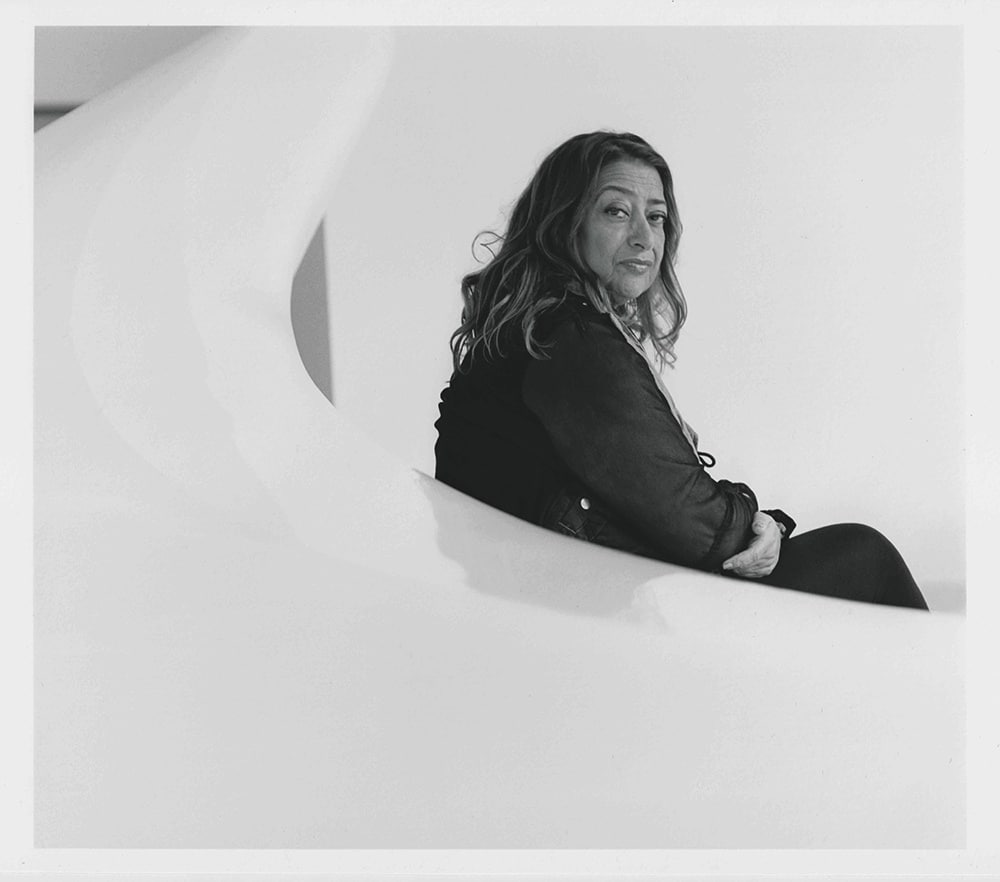 A photo of Zaha Hadid sitting on a slick white surface. She is looking back at the camera over her shoulder.